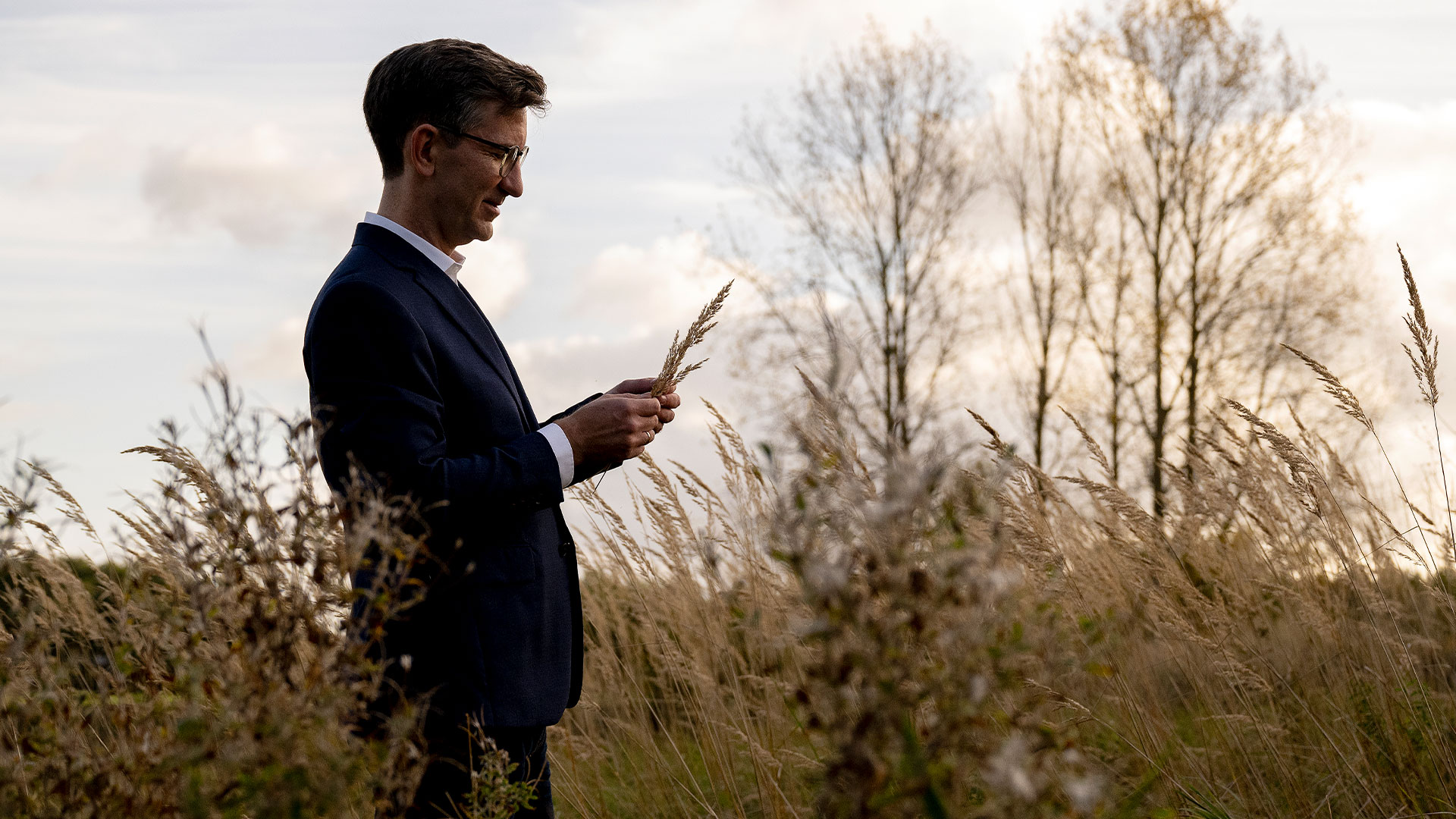 Man standing in tall grasses looking at a sample that he is holding in his hand.